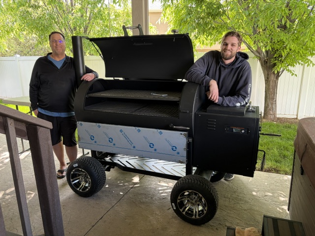 Dan Moe Nelson and Tyler Nelson standing with the Mo'Licious smoker